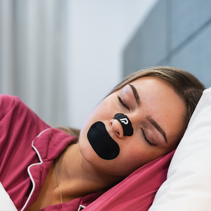 Woman in pink pyjamas sleeping with a black nasal strip and black mouth tape