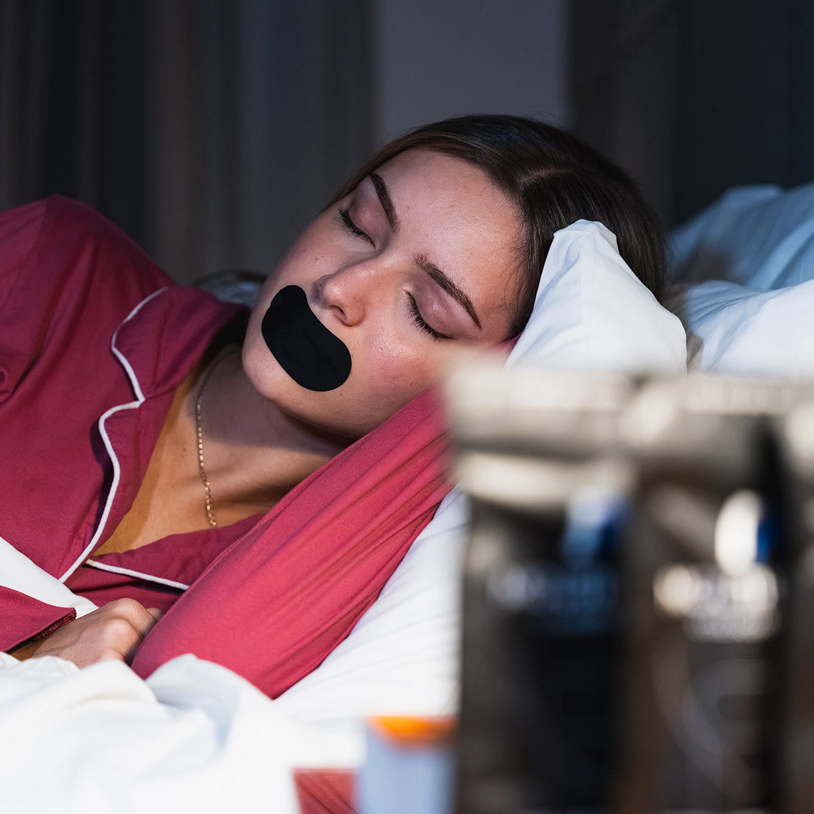Woman lying in bed wearing black mouth tape beside blurred bedside items